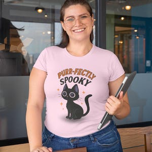 Może przedstawiać: Jasnor&oacute;żowa koszulka z napisem "Purr-fectly Spooky" i czarną grafiką kota. Kot ma duże oczy i siedzi ze zwiniętym ogonem. Koszulka jest noszona z niebieskimi dżinsami.