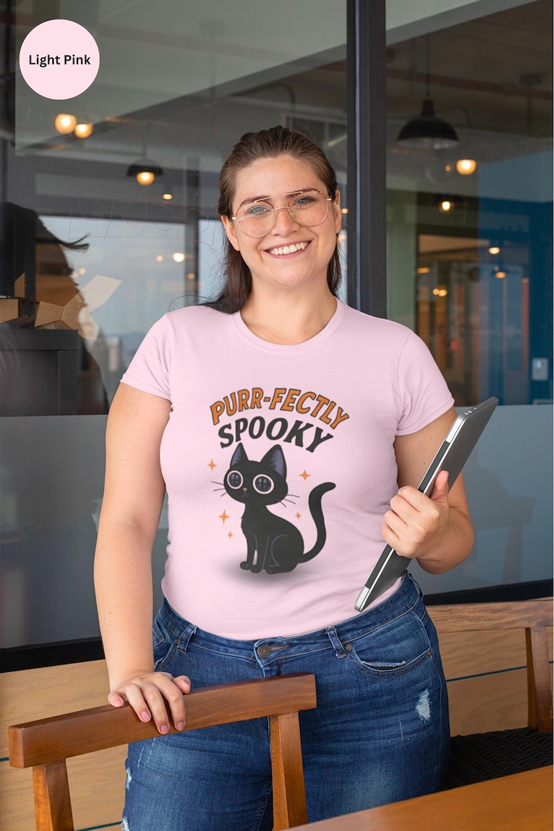 Może przedstawiać: Jasnor&oacute;żowa koszulka z napisem "Purr-fectly Spooky" i czarną grafiką kota. Kot ma duże oczy i siedzi ze zwiniętym ogonem. Koszulka jest noszona z niebieskimi dżinsami.