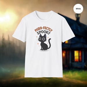 Może przedstawiać: Biała koszulka z kresk&oacute;wkowym czarnym kotem i napisem "Purr-fectly Spooky". Kot ma duże oczy i siedzi ze zwiniętym ogonem. Projekt zawiera pomarańczowe akcenty gwiazd.