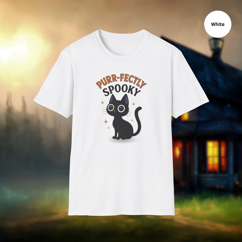 Może przedstawiać: Biała koszulka z kresk&oacute;wkowym czarnym kotem i napisem "Purr-fectly Spooky". Kot ma duże oczy i siedzi ze zwiniętym ogonem. Projekt zawiera pomarańczowe akcenty gwiazd.