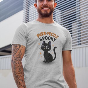Może przedstawiać: Szara koszulka sportowa z ilustracją czarnego kota z kresk&oacute;wki i napisem "Purr-fectly Spooky" w kolorze pomarańczowym i czarnym. Kot ma duże oczy i siedzi. Koszulka jest w kolorze jasnoszarym.
