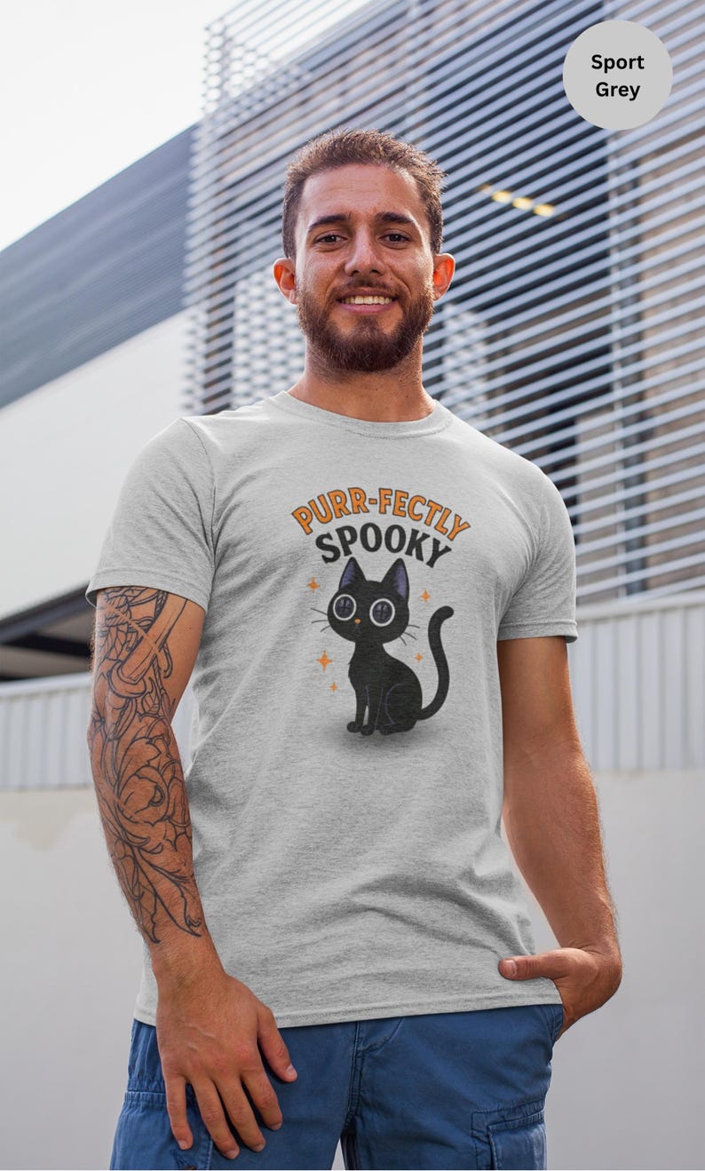 Może przedstawiać: Szara koszulka sportowa z ilustracją czarnego kota z kresk&oacute;wki i napisem "Purr-fectly Spooky" w kolorze pomarańczowym i czarnym. Kot ma duże oczy i siedzi. Koszulka jest w kolorze jasnoszarym.