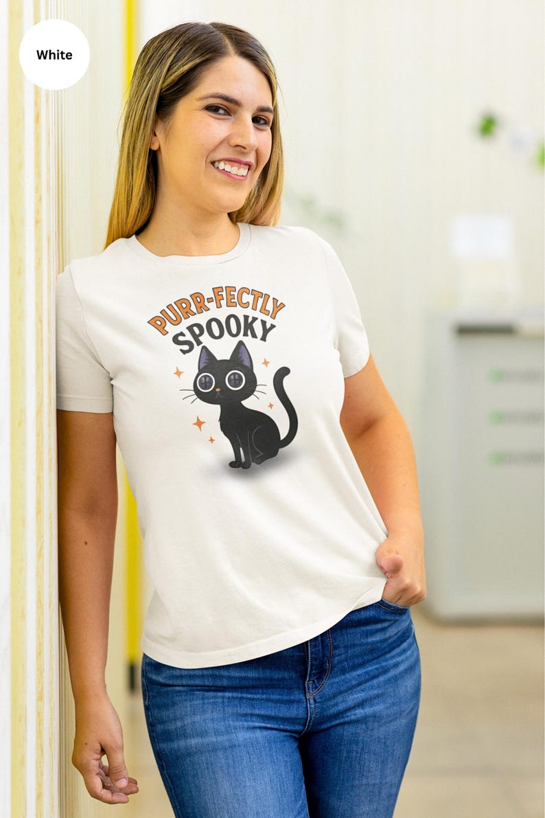 May include: White t-shirt with a cartoon black cat illustration and the text "Purr-fectly Spooky" in orange and black. The cat has button eyes and is surrounded by orange stars. The shirt is paired with blue jeans.