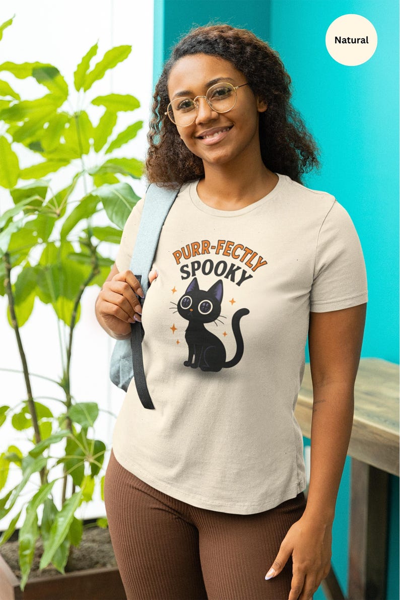 Może przedstawiać: Kremowa koszulka z napisem "Purr-fectly Spooky" i ilustracją czarnego kota. Kot ma duże białe oczy i siedzi ze zwiniętym ogonem. Koszulka jest zestawiona z brązowymi spodniami.