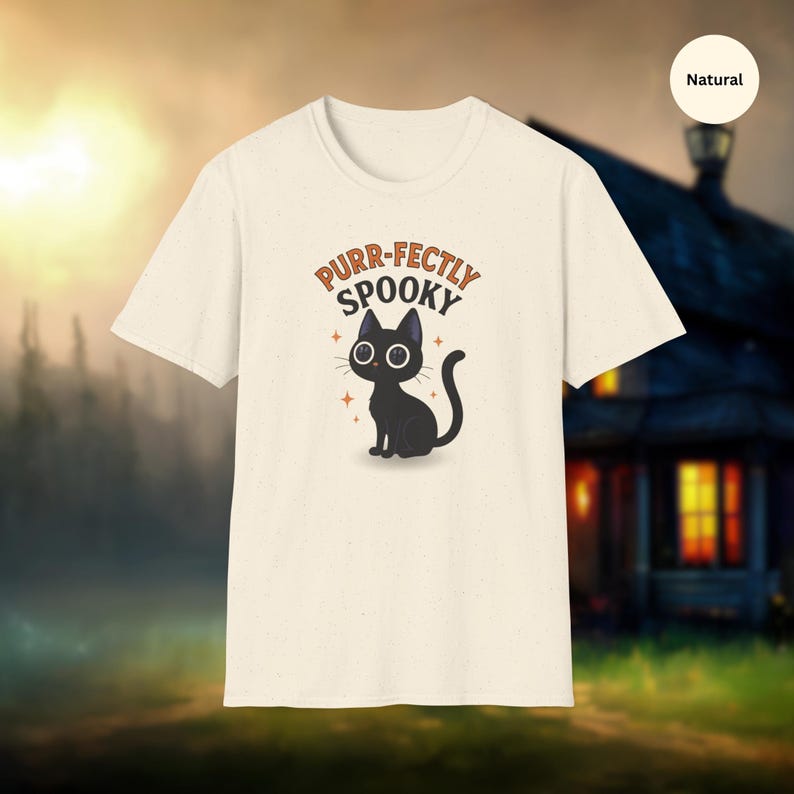 Może przedstawiać: Kremowa koszulka z ilustracją czarnego kota i napisem "Purr-fectly Spooky" w kolorze pomarańczowym i czarnym. Projekt zawiera pomarańczowe akcenty w postaci gwiazd. Idealna na Halloween.