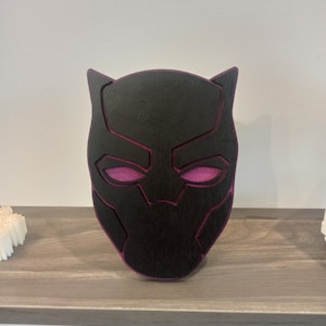 Puede incluir: Una máscara de madera negra con contorno y detalles de ojos morados, que se asemeja a Black Panther. La máscara se exhibe en una estantería de madera, mostrando un artículo temático de superhéroes.