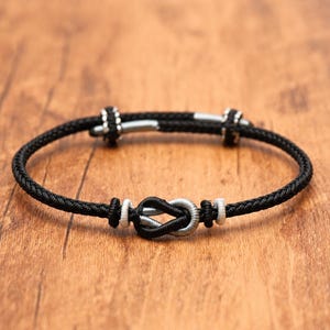 May include: A black cord bracelet featuring a central infinity-style knot design with silver-toned accents. The bracelet includes two adjustable sliding silver-toned metal beads on the cord, allowing for a flexible fit around the wrist.
