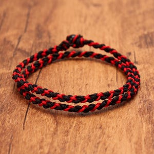 May include: A double-wrap bracelet constructed from braided black and red cord. The design features a continuous twisted pattern with a small, dark, rounded knot closure at the top, creating a circular shape with two distinct strands of cord.