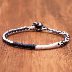 May include: A bracelet constructed from braided navy blue and white cord, featuring a circular knot closure. The design includes a section wrapped in solid navy blue thread and a section wrapped in solid white thread, creating a contrast.