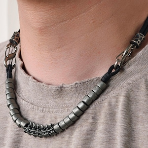 May include: A silver-toned necklace with a black cord. The necklace features cylindrical and rectangular metal beads, connected by a clasp. The design is visible against a neutral-colored shirt.