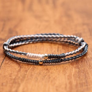 May include: A double-strand bracelet featuring dark gray and white braided cord. One strand includes a white knotted section and a small metallic gold bead, while the second strand is a solid dark gray cord accented with a single metallic gold bead.