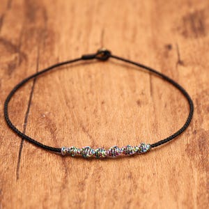 May include: A thin black cord bracelet featuring a central row of six small, textured beads with a multicolored, iridescent finish. The bracelet is secured with a simple sliding knot closure, creating a minimalist and adjustable design.