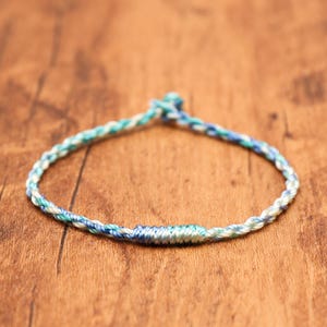 May include: A thin, braided cord bracelet featuring a variegated color palette of light blue, turquoise, and white threads. The bracelet is constructed from twisted strands and includes a small, tightly wrapped section of thread near the center.