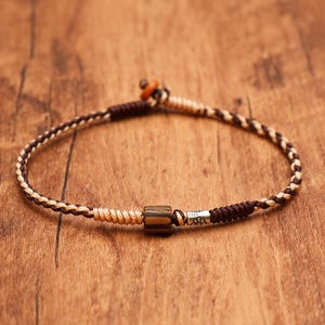May include: A braided cord bracelet featuring a mix of dark brown and beige threads. The design includes a central cylindrical striped bead, a small silver-toned metal accent piece, and a sliding knot closure with a small round bead at the end.