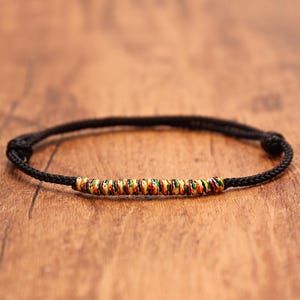 May include: A black cord bracelet featuring a central cylindrical section wrapped in thin, multicolored thread in shades of red, yellow, and green. The bracelet includes two adjustable sliding knots on either side of the central decorative element.