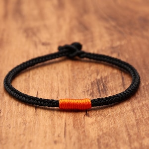 May include: A thin black cord bracelet featuring a single, vibrant orange thread accent wrapped around a section of the band. The bracelet is constructed from braided material and includes a small, simple knot closure at the top of the circular loop.
