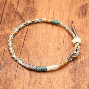 May include: A braided cord bracelet featuring a mix of cream, sage green, and metallic gold threads. The design includes sections of tightly wrapped thread in grey, green, cream, and gold, with a small sliding knot closure and metal accents.