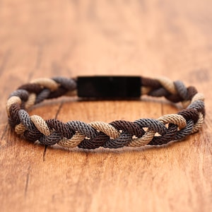 May include: A braided bracelet featuring three interwoven cords in shades of tan, dark brown, and charcoal gray. The bracelet is secured with a smooth, rectangular black metal clasp that connects the ends of the braided cord design.