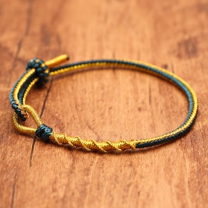 May include: A thin bracelet constructed from braided dark teal and metallic gold cord. The design features a twisted gold section, a small loop closure, and a sliding knot mechanism with a gold-toned cylindrical end piece for adjustment.