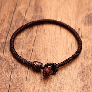 May include: A dark brown braided cord bracelet featuring a cylindrical wooden bead with a white cloud engraving and a small, round, textured reddish-brown wooden bead acting as a toggle closure for the adjustable knotted cord loop design.