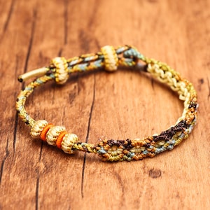 May include: A woven cord bracelet featuring a complex pattern of brown, tan, and light blue threads. The bracelet includes three metallic gold-toned beads separated by two bright orange rings, along with adjustable sliding knots in tan.