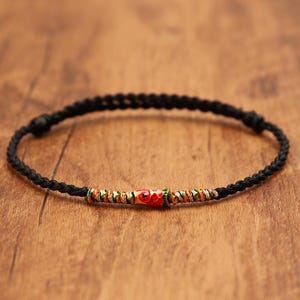 May include: A black braided cord bracelet featuring a central cylindrical bead. The bead is decorated with a vibrant red, green, and gold pattern. The bracelet includes adjustable sliding knots on each side of the central decorative element.