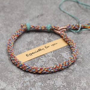 May include: A braided bracelet made of multi-colored thread in shades of red, yellow, blue, and green. The bracelet features a light blue sliding knot closure and a tassel end. A tan rectangular paper tag is tucked inside with Especially for you.
