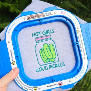 Puede incluir: Una camiseta gris con un diseño bordado de un tarro de pepinillos y el texto "HOT GIRLS LOVE PICKLES". La camiseta se sujeta en un bastidor de bordado azul. El bastidor tiene advertencias de seguridad y el texto "Made in the USA".