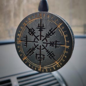 Op de afbeelding: Een zwarte houten autohanger met een gouden kompasontwerp en runen rond de rand. De hanger hangt aan een zwart touwtje.
