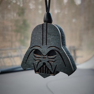Può includere: Un ciondolo per auto in legno nero a forma di testa di Darth Vader. Il ciondolo è appeso a uno stringa nera.