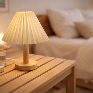 Peut inclure: Une petite lampe avec un abat-jour plissé crème est posée sur un socle en bois, diffusant une lumière chaleureuse. La lampe est sur une table de chevet en bois clair, à côté d'un verre d'eau et d'une pile de livres. En arrière-plan, un lit avec une literie blanche.