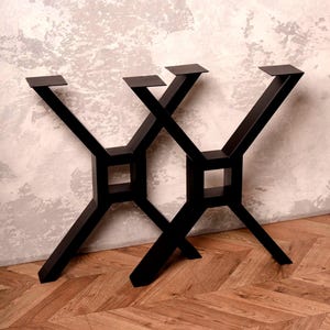 May include: Two black metal table legs with a geometric X-shaped design. Each leg has a square base and a flat top for attaching a tabletop. The legs are made of solid metal and are suitable for a modern or industrial style table.