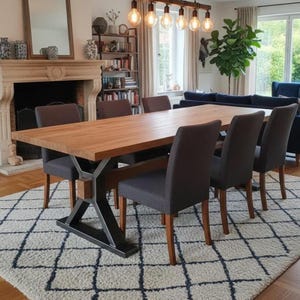 May include: A wooden dining table with a light brown finish and a metal base, surrounded by six grey upholstered chairs. The table is set on a patterned rug. The room has a fireplace and a large window.