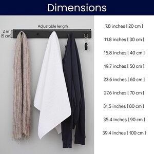 May include: A black metal wall-mounted coat rack with three items hanging from it: a beige scarf, a white towel, and a navy blue sweatshirt. The dimensions are listed in inches and centimeters.