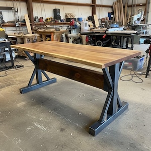 Puede incluir: Una mesa de madera grande con una tapa de color marrón claro y una viga de soporte marrón oscuro. La mesa tiene patas de metal negro con un diseño en forma de X. La mesa está en un taller.