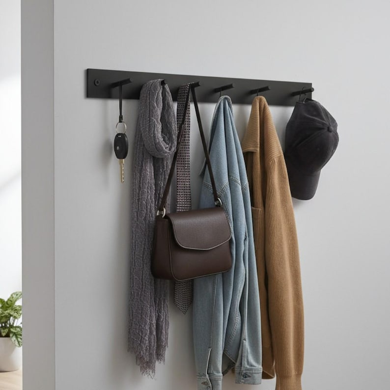 May include: A black wall-mounted coat rack with six hooks. Items hanging on the hooks include a set of keys, a gray scarf, a brown purse, a patterned tie, a denim jacket, a tan sweater, and a black baseball cap.