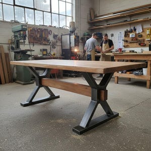 Op de afbeelding: Een grote houten tafel met een lichtbruin tafelblad en donkere metalen poten. De poten hebben een X-vormig ontwerp en de tafel staat in een werkplaats met gereedschap en twee personen op de achtergrond.