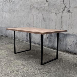 Könnte beinhalten: Ein rechteckiger Holztisch mit hellbrauner Oberfläche. Der Tisch hat schwarze Metallbeine. Die Beine sind ein schlichtes, modernes Design. Der Tisch steht vor einer grauen Betonmauer.