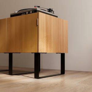 May include: A light oak wood cabinet with two doors and a small silver handle, supported by black metal U-shaped legs. A black turntable with a silver tone arm and red cartridge is positioned on the flat top surface of the wooden cabinet.