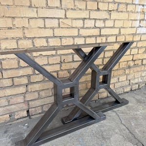 Può includere: Un tavolo consolle in metallo con base geometrica. La base è in metallo grigio scuro e presenta un design a forma di X con accenti quadrati. Il piano del tavolo non è visibile. Il tavolo è progettato per essere posizionato contro un muro.