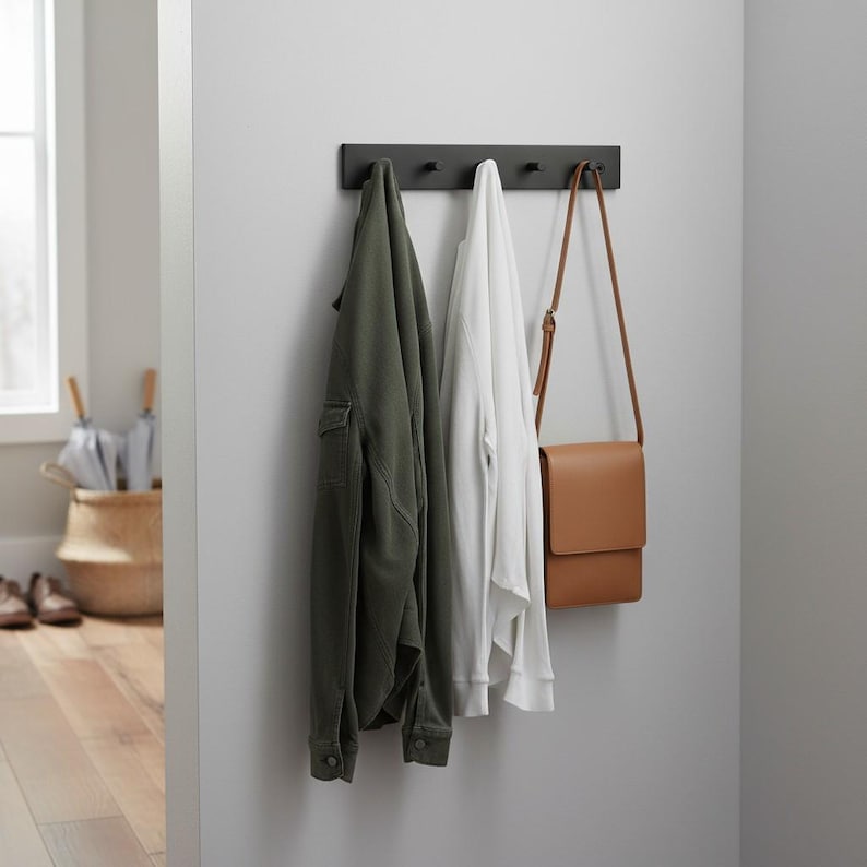 May include: A dark gray wall-mounted coat rack with four hooks. An olive green jacket, a white sweatshirt, and a brown leather crossbody bag hang from the hooks. The items are displayed against a light gray wall.