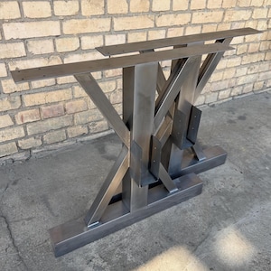 Puede incluir: Un par de patas de mesa de acero sin terminar. Las patas tienen un diseño en forma de X con una base rectangular y una parte superior plana con agujeros pretaladrados para fijar una mesa. El metal tiene un aspecto crudo e industrial.