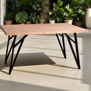 Puede incluir: Mesa rectangular de madera con superficie marrón claro y patas oscuras con diseño geométrico. La mesa está situada al aire libre, con vegetación de fondo.