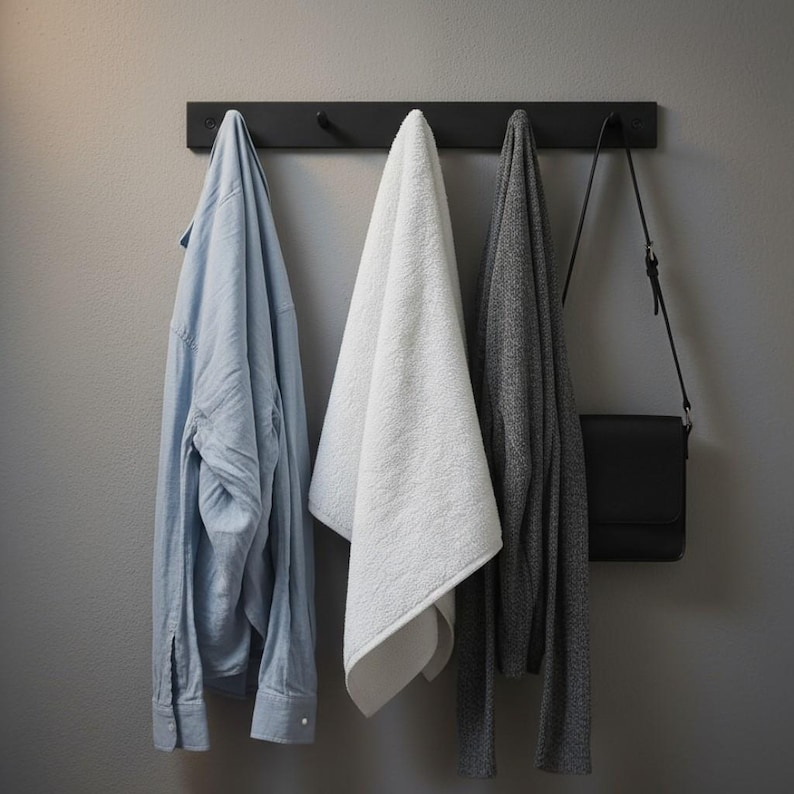 May include: A black wall-mounted coat rack with four hooks holds a light blue button-down shirt, a white towel, a gray sweater, and a black crossbody bag. The items hang against a gray wall.