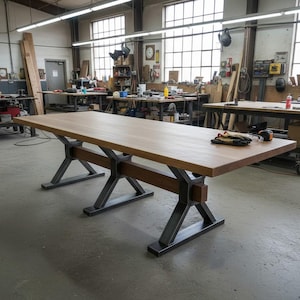Puede incluir: Una mesa rectangular de madera clara. La mesa está sostenida por patas de metal oscuro con diseño en forma de X. La mesa se encuentra en un taller, probablemente para la fabricación de muebles.