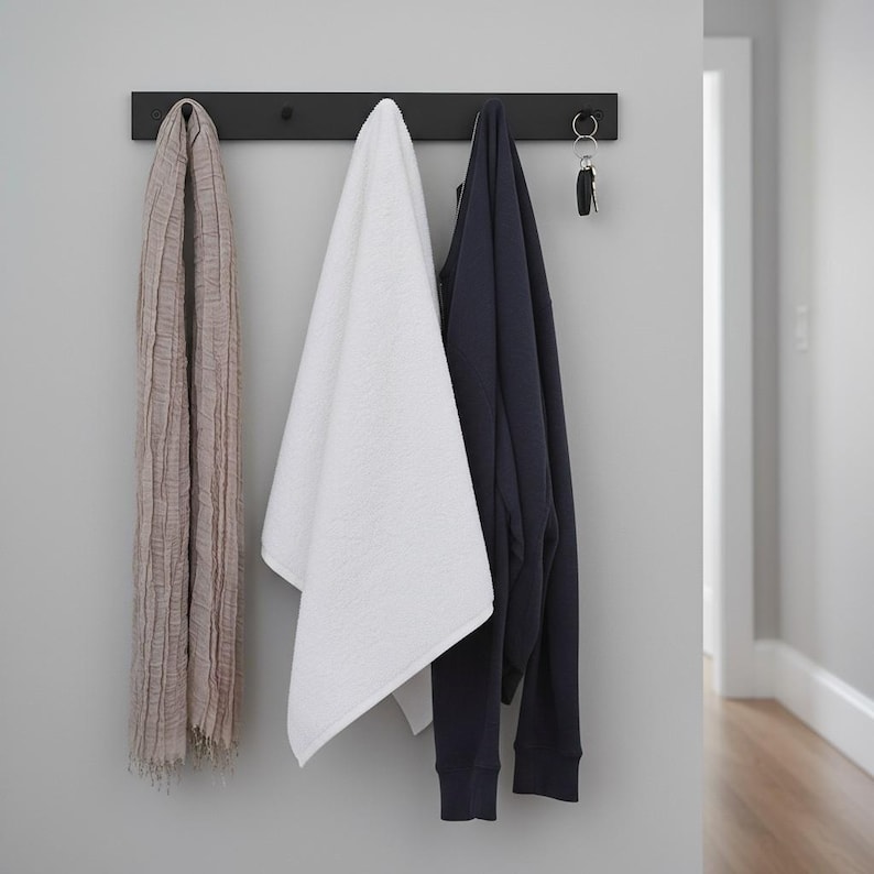 May include: A black wall-mounted coat rack with a scarf, a white towel, a navy blue sweatshirt, and keys hanging from it. The scarf is a light brown color, and the towel is white. The sweatshirt is a solid navy blue color.