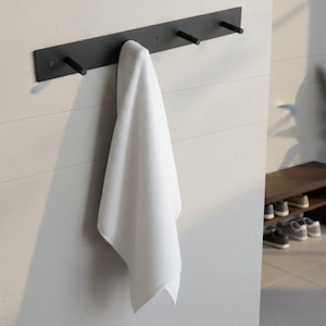May include: A black metal wall-mounted towel rack with three pegs, holding a folded white towel. The rack is mounted on a white wall with horizontal lines. The design is minimalist and modern, suitable for a bathroom or spa.