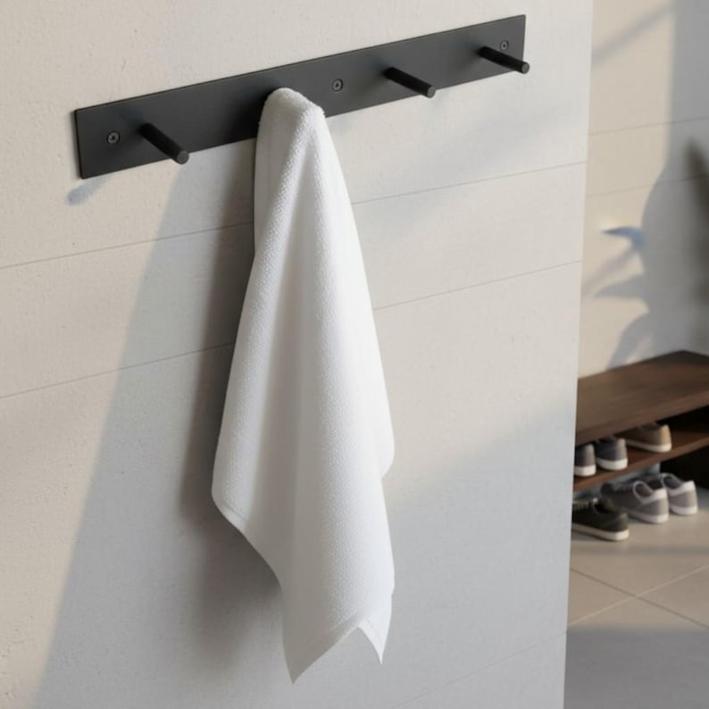 May include: A black metal wall-mounted towel rack with three pegs, holding a folded white towel. The rack is mounted on a white wall with horizontal lines. The design is minimalist and modern, suitable for a bathroom or spa.