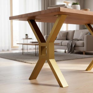 May include: A wooden dining table with a light brown tabletop and gold-coloured metal legs. The legs have an X-shaped design. The table is in a living room setting.