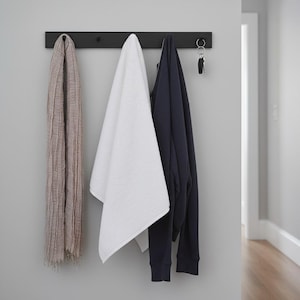 May include: A black wall-mounted coat rack with a scarf, a white towel, a navy blue sweatshirt, and keys hanging from it. The scarf is a light brown color, and the towel is white. The sweatshirt is a solid navy blue color.
